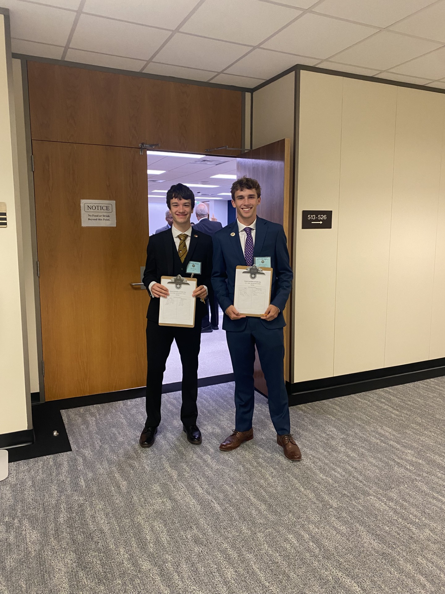 Two pages standing in front of a committee room holding up clipboards with bill documents.